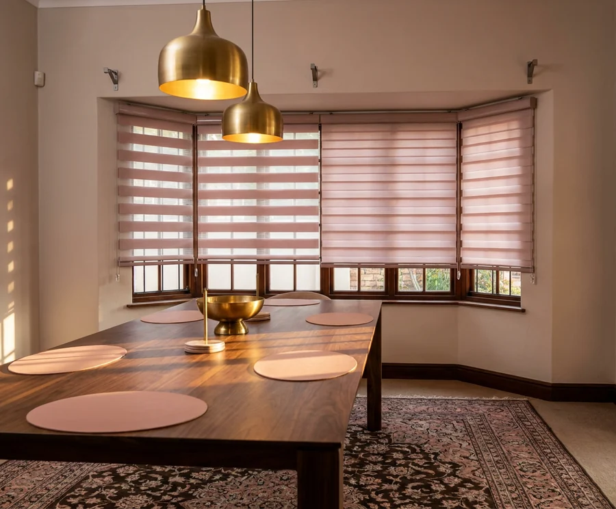 Day/night blinds across the three panes of a Melrose dining-room bay window, bands part-aligned for view and part-closed for privacy under brass pendants